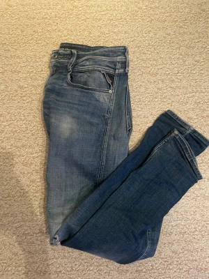 Replay Anbass jeans blå 33/32 - Snygga Replay Anbass jeans i klassisk blå tvätt med lätt slitna detaljer. Modellen har smal passform och raka ben, med typiska Replay-sömmar och fem fickor. Perfekt för dig som gillar stilrena och moderna jeans.
