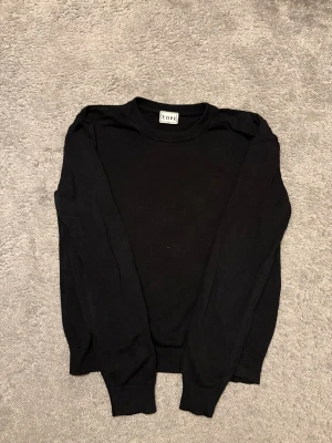 Svart långärmad stickad tröja från COPE - En stilren svart långärmad stickad tröja från COPE i klassisk modell. Tröjan har rund hals och ribbade muddar vid ärmslut och nederkant. Perfekt för dig som gillar enkel och clean stil. Materialet känns mjukt och bekvämt mot huden. Och perfekt om du vill ha lite extra stil i vardagen.  