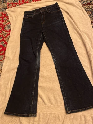 Bootcut mörkblå jeansbyxor - Snygga mörkblå jeansbyxor med klassisk bootcut-passform och fem fickor. Jeansen har en rak midja och är tillverkade i slitstarkt denim-material. Perfekta för dig som gillar en retro vibe och vill ha ett par jeans med lite extra vidd nedtill.