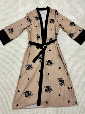 Beige abaya med svarta broderier och bälte - Lång beige abaya med svarta broderade blommor och svarta detaljer vid krage och ärmslut. Kappan har ett matchande bälte med metallspänne i midjan och trekvartsärmar. Snygg och unik design som sticker ut.