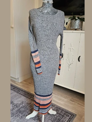 Ribbad långklänning med färgglada ränder - Ribbad långklänning i grått med färgglada ränder i orange, marinblå, rosa och vitt på ärmar och nederkant. Klänningen har lång ärm och figurnära passform. 