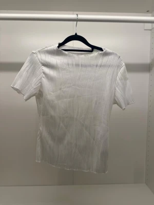 Vit ribbad blus med struktur - Säljer en vit blus med korta ärmar och snyggt ribbad struktur i tyget. Toppen har en rak passform och rund halsringning. Materialet är mjukt och har en lätt glans, vilket ger en fräsch och modern vibe. Perfekt för dig som gillar stilrena plagg med lite extra detalj.