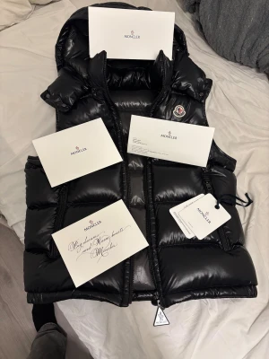 Svart dunväst från Moncler med huva - Säljer en svart, glansig dunväst från Moncler i modell Bormes Gilet, storlek M/L. Skick 10/10 nästan aldrig använd, hör av er vid funderingar. ÄKTA kontakta för mer bevis 😁