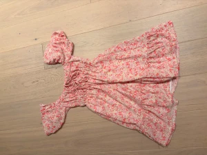 Blommig kortklänning från Gina Tricot - Supersöt kortklänning från Gina Tricot med rosa och vita blommor, puffiga kortärmar och smockad midja med snörning framtill. Lätt och luftig, perfekt för varma dagar. Materialet känns som bomull eller polyester. Aldrig använd 