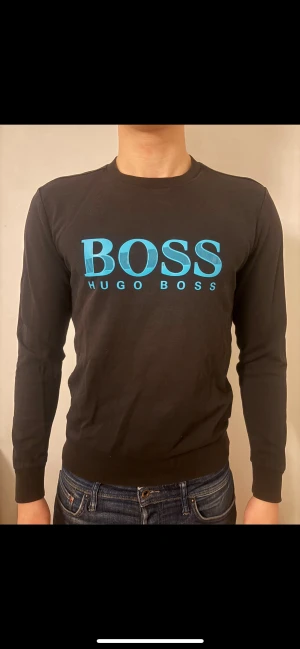 Svart långärmad tröja Hugo Boss - Svart långärmad tröja från Hugo Boss med stort blått BOSS-tryck på bröstet. Tröjan har rund hals, ribbade muddar och är tillverkad i mjuk bomull. Snygg och enkel design som passar perfekt till jeans.