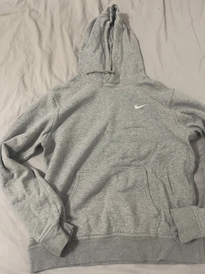 Grå hoodie från Nike med huva - Grå Nike hoodie med klassisk logga på bröstet. Tröjan har huva med snörning och en stor magficka framtill. Tillverkad i mjuk bomullsmix, perfekt för chill eller träning. Långärmad och loose passform för skön vibe.