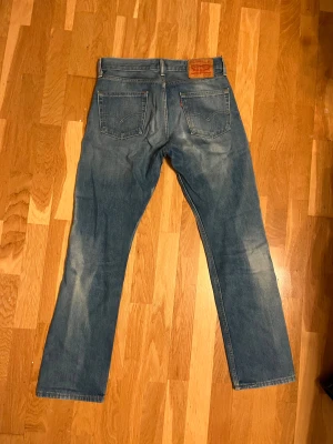 Levi's 501 blå jeans straight fit - Klassiska Levi's 501 jeans i blå tvätt med snygga slitningar och raka ben. Modellen har fem fickor, knappgylf och den ikoniska läderpatchen bak i midjan. Perfekt för dig som gillar tidlös stil och vill ha ett par jeans med autentisk känsla.