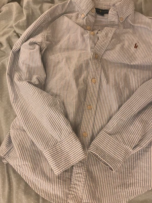 Randig skjorta från Ralph Lauren - Snygg skjorta från Ralph Lauren med klassiskt blåvitt randigt mönster och broderad logga på bröstet. Skjortan har lång ärm, knappar framtill och krage med knapp. Tillverkad i mjuk bomull som känns skön mot huden. Perfekt för dig som gillar stilrena och tidlösa plagg. Priset kan diskuteras.