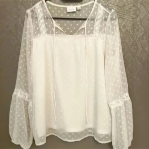 Vit prickig blus från Vila - Superfin vit blus från Vila med transparenta, långärmade ärmar i mesh och små prickar. Blusen har spetsdetaljer vid axlar och ärmar, v-ringning med knytband och en luftig, flowy passform. Perfekt till jeans eller kjol för en fräsch look.