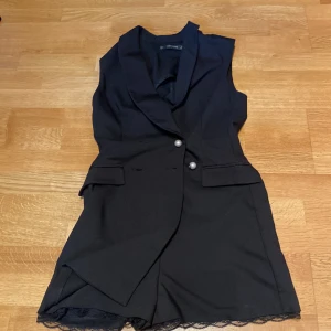 Zara Basic - Svart ärmlös kavajdräkt  - Svart ärmlös kavajdräkt från Zara Basic med snygga pärlknappar framtill och spetsdetaljer vid bensluten. Dräkten har klassisk kavajkrage, fickor och ett bälte baktill för extra stilpoäng. Perfekt för dig som vill sticka ut med en modern twist på kostymlooken.