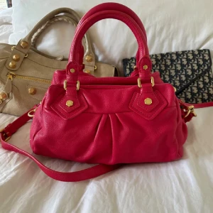 Marc Jacobs väska - Cool röd handväska i skinn från Marc by Marc Jacobs med guldiga metalldetaljer och dragkedja. Väskan har både handtag och axelrem samt ett snyggt innerfoder med logotypmönster. Perfekt storlek för att få plats med allt du behöver. 