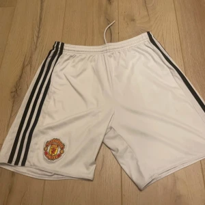Manchester United shorts Adidas S - Snygga vita Manchester United shorts från Adidas med svarta ränder längs sidorna och klubbens emblem på ena benet. De har elastisk midja med snörning och är gjorda i ett lätt och skönt polyestermaterial, perfekt för fotbollsträning eller match.