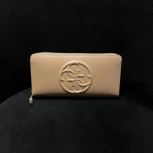 Beige plånbok från Guess i skinn - Stilren beige plånbok från Guess i skinn med präglad logga på framsidan. Plånboken har flera fack, dragkedja runt om och detaljer i guld. Perfekt för att hålla ordning på kort och kontanter.