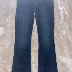 True Religion Becca Bootcut Jeans - Mörkblå jeans från True Religion, modell Becca Mid Rise Bootcut. Snygg passform med bootcut-ben. Klassiska bakfickor med dekorativa sömmar och orange detaljer vid knappar och sömmar. Tillverkade i stretchig denim för extra komfort. Använda 5 gånger