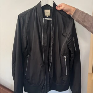 Svart bomberjacka Jack & Jones Premium  - Svart bomberjacka från Jack & Jones Premium med silvriga dragkedjor, ribbade muddar och klassisk krage. Jackan har flera fickor med dragkedja och är tillverkad i ett lätt, syntetiskt material. Perfekt för dig som gillar en clean och stilren look.