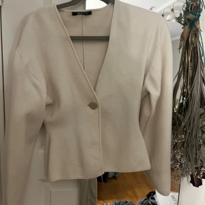 Beige kofta med v-ringning från Gina Tricot - Snygg beige kofta från Gina Tricot med v-ringad hals och en guldfärgad knapp framtill. Modellen är lite croppad och har långa ärmar. Den är i storlek S och säljer då den inte kommer till användning.