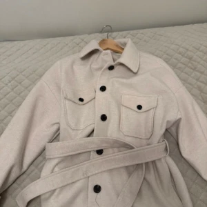 Beige overshirt med bälte från NLY Trend - Snygg beige overshirt från NLY Trend med bälte i midjan och svarta knappar. Jackan har två bröstfickor med lock och klassisk krage. Perfekt för lager-på-lager och en clean look.
