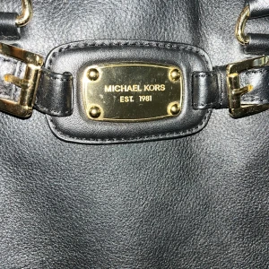 Svart axelväska från Michael Kors - Snygg svart axelväska från Michael Kors med guldfärgade kedjedetaljer och metallplatta framtill. Väskan är tillverkad i skinn och har både handtag och axelrem, vilket gör den enkel att bära. Perfekt för dig som gillar stilrena accessoarer med lyxig känsla.