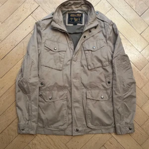 Beige vindjacka från Woolrich - Snygg beige vindjacka från Woolrich med flera praktiska fickor framtill och axelparti med knappdetaljer. Jackan har hög krage, dragkedja och tryckknappar. Perfekt för dig som gillar en stilren och funktionell look.
