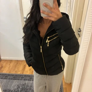 Svart pufferjacka med guldiga dragkedjor - INTRESSEKOLL! Snygg svart pufferjacka från Hollies med guldiga dragkedjor och coolt leopardmönstrat foder. Jackan har huva, flera fickor med dragkedja och en figurnära passform. Köpt för 3299kr på Bubblerooom. 