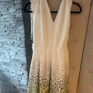 Vit leopardmönstrad klänning Samofono - Supersnygg vit kortklänning från Samofono med v-ringning och leopardmönster i grått och grönt nertill. Klänningen har omlott-detalj i ryggen och knytband i midjan. Perfekt för dig som vill sticka ut med en cool och fräsch look.