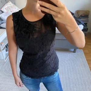 Svart spetslinne med blommor - Supersnygg svart ärmlös linnetopp med transparent mesh upptill och dekorativa tygblommor vid halsringningen. Toppen har spetsdetaljer över hela framsidan och en feminin, figurnära passform. Perfekt för dig som gillar unika detaljer och romantisk stil.
