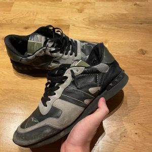 Valentino Rockrunner sneakers camo - Snygga Valentino Rockrunner sneakers i svart, grått och olivgrönt med camouflagemönster. Skorna har svarta snören, platt sula och detaljer i mocka och skinn. Ikoniska nitar på hälen och cool streetstil. Perfekta för dig som gillar exklusiva sneakers med edge. Har tyvärr en liten defekt på hälen då jag har trampat ner dem lite på innersidan men det är inget man känner och det syns knappt. Dustbags ingår!