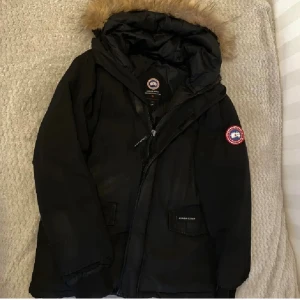 Svart dunjacka från Canada Goose - Säljer en svart dunjacka från Canada Goose med klassisk patch på ärmen och huva med fuskpäls. Jackan har stora fickor framtill, dragkedja och kardborreknäppning. Perfekt för kalla vinterdagar och riktigt snygg streetstil.