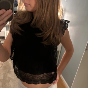 Svart spetsblus med volangdetaljer - Supersnygg svart blus i spets med fina volangdetaljer Blusen är ärmlös och har ett vackert mönster i spetsen som ger en elegant känsla. Använd några få gånger❤️❤️