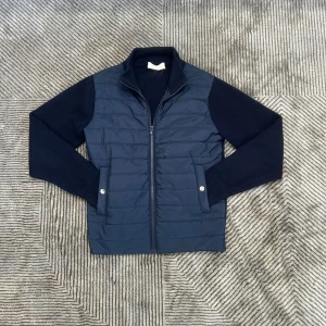 Mörkblå cardigan från Sayless - Snygg mörkblå pufferjacka från Sayless med stickade ärmar och quiltad front. Jackan har dragkedja framtill, ståkrage och två fickor med tryckknappar. Perfekt för dig som gillar en sportig och stilren look.