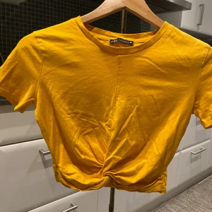 Gul croppad topp från Zara - Snygg gul croppad topp från Zara med korta ärmar och twistad detalj framtill vid midjan. T-shirten har rund halsringning och är tillverkad i mjuk bomull, perfekt för en färgstark look.