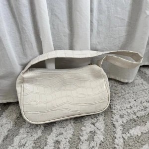 Vit axelväska i croco-imitation - Snygg vit axelväska med croco-mönstrad yta och glansig finish. Väskan har en bred axelrem och stängs med dragkedja i guld. Perfekt storlek för det viktigaste och ger en trendig touch till din outfit.