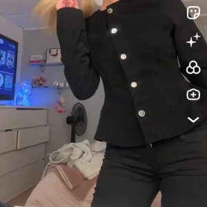 Jeans topp från ginatricot  - Snygg svart jeanstopp med raka linjer och markerad midja. Kavajen har stora silverfärgade knappar framtill som ger en clean och stilren look. Perfekt att styla med både jeans och kostymbyxor. Klassisk krage och långärmad modell.