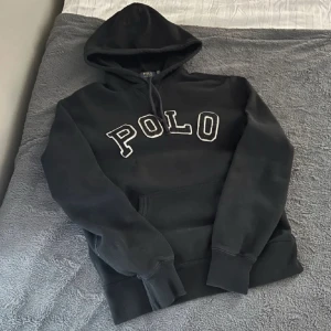 Svart hoodie från Polo Ralph Lauren - Svart hoodie från Polo Ralph Lauren med stor broderad POLO-text i vitt på bröstet. Klassisk huva tröja, har dock tagit bort snörert. Perfekt för dig som gillar en clean stil!