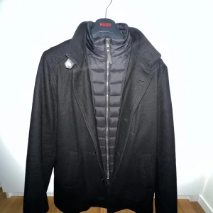 Svart rock från Rockandblue, stl 54 - Säljer en svart rock från Rockandblue med snygg krage och avtagbar quiltad innerdel med dragkedja. Jackan har knappar framtill, fickor på sidorna och en bröstficka med dragkedja. Perfekt för kyliga dagar och ger en clean look.