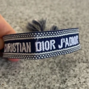 Blått Christian Dior J'Adior armband - Säljer ett blått armband från Christian Dior med texten 'J'Adior' broderad i vitt. Armbandet är gjort i tyg och har snygga tofsar i ändarna för en cool och trendig look. Perfekt accessoar för att lyfta din outfit.