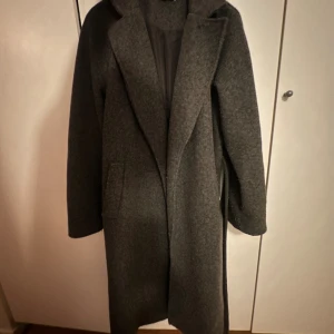 Mörkgrå ullkappa med bälte - Säljer en mörkgrå kappa i ull med krage och bälte i midjan. Kappan har lång ärm, rak passform och två fickor framtill. Perfekt för dig som vill ha något varmt och snyggt till höst och vinter🩶Jag säljer då den inte längre kommer till användning. OBS den har nopprat vilket visas i sista 2 bilderna! 