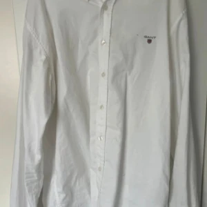 Vit skjorta från Gant, 176 cm - Klassisk vit skjorta från Gant med broderad logga på bröstet. Skjortan har lång ärm, knappar framtill och är tillverkad i bomull. Perfekt för en clean och stilren look.