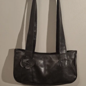Svart axelväska  med blomdetalj - Stilren svart axelväska  med lång rem och en söt blomdetalj framtill. Väskan har en avlång form och stängs med dragkedja upptill. Perfekt för dig som gillar minimalistisk design med en liten twist. Har två små skavor men inget som syns så mycket 