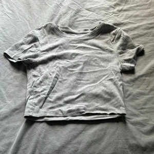Grå magtröja - En grå ribbad topp från New Yorker i storlek S. Modellen är ribbad och croppad med korta ärmar och rund halsringning. 
