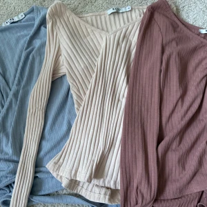 Tre ribbade långärmade toppar från NA-KD - Säljer tre snygga ribbade toppar från NA-KD i färgerna ljusblå, beige och mörkrosa. Två har v-ringning och lång ärm, den ljusblå är omlott med knyt i sidan. Perfekta för dig som gillar stilrena och bekväma plagg med lite edge.
