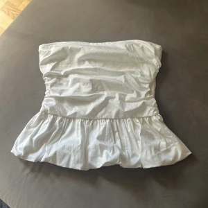 Vit volang tubtopp från Zara - Säljer en vit tubtopp från Zara i storlek M. Toppen har en snygg volang nedtill och är axelbandslös med en lätt rynkad effekt över bröstet. Perfekt för dig som gillar en clean och fräsch look med lite extra volym. Materialet känns mjukt och luftigt.