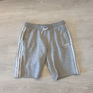 Grå Adidas shorts med vita ränder - Snygga grå shorts från Adidas med klassiska vita ränder längs sidorna och logga framtill. De har snörning i midjan och är gjorda i mjuk bomullsmix, perfekta för chill eller träning. Fickor på sidorna för mobilen eller nycklarna.