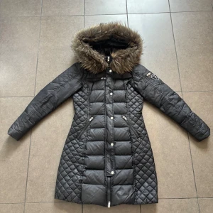 Svart lång dunjacka Rockandblue - Snygg svart lång dunjacka från Rockandblue med quiltade detaljer och pälsimitation på huvan. Jackan har flera dragkedjor, knappar och en broderad logga på ärmen. Figurnära passform och långa ärmar, perfekt för kalla dagar.