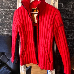 Röd quiltad jacka från A-One, stl S - Snygg röd quiltad jacka från A-One med svarta detaljer längs sidor och rygg. Jackan har dragkedja framtill, ståkrage och praktiska fickor. Vertikala sömmar ger en sportig look och ärmarna har dragkedjor för extra stilpoäng.