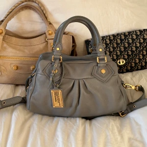 Grå Q Classic Baby Groove väska! - Snyggaste ljusgrå handväskan i äkta läder från Marc by Marc Jacobs. Den ikoniska modellen Q Classic Baby Groove! En populär och eftertraktad vintagemodell med gulddetaljer och dubbla handtag. Långa bandet finns och är i gott skick. Väskan är i nyskick. Lädret är helt utan repor eller hål. Finns två mindre fläckar (se bild) men märks knappt. Skriv privat för fler bilder🤍
