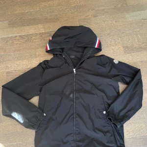 Svart vindjacka från Moncler med huva - Svart vindjacka från Moncler med huva och dragkedja. Jackan har Moncler-logga på ärmen och snygga röd-vita-blå detaljer på huvan. Tillverkad i polyester och har elastiska muddar vid ärmslut. Perfekt för dig som vill ha en stilren och sportig look.