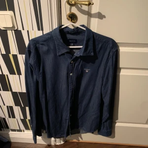 Mörkblå skjorta från GANT, storlek L - Snygg mörkblå skjorta från GANT i storlek L med klassisk krage och knäppning framtill. Skjortan har lång ärm och diskret GANT-logga broderad på bröstet. Perfekt för en clean och stilren look.