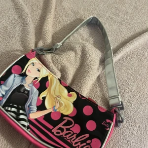 Barbie axelväska med rosa prickar - Snygg axelväska från Barbie med silverfärgat axelband och rosa detaljer. Framsidan har ett tryck med Barbie och stora rosa prickar på svart bakgrund, medan baksidan är helrosa med glittrigt Barbie-logo. Insidan är svart med dragkedja och hjärtformad zip-pull. Perfekt för dig som gillar färg och Barbie-vibe.