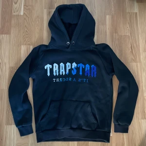 Trapstar Hoodie - Svart hoodie ifrån Trapstar med stor logga i blått framtill. Klassisk känguruficka, huva med metallöljetter och mjukt material. Kolla bild 3 för ända synliga defekt. 